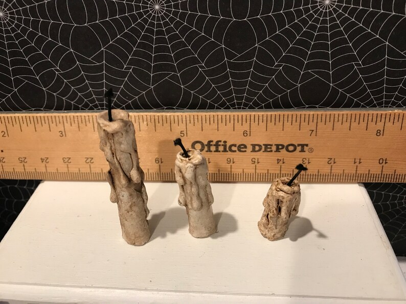 Puede incluir: Tres velas de cera blanca, goteando, con mechas negras. Las velas est&aacute;n dispuestas en fila sobre una superficie blanca. Una regla es visible en el fondo con el texto "Office DEPOT" impreso en ella.