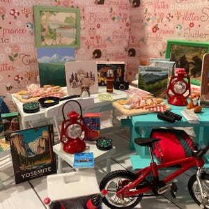 May include: A miniature camping scene with a red bicycle, picnic tables, and camping-themed decor. Includes a red lantern, small paintings, and a fox figurine. The background features a floral wallpaper.