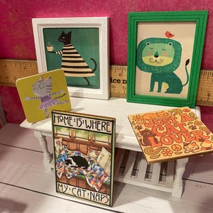 K&ouml;nnte beinhalten: Eine Sammlung von Miniatur-Wandkunst und quadratischen Schildern mit Katzen- und L&ouml;wenillustrationen. Ein Schild tr&auml;gt die Aufschrift "HOME IS WHERE MY CAT NAPS" und ein anderes "JUST A CAT LOVER". Ein Holzlineal dient als Gr&ouml;&szlig;enreferenz.