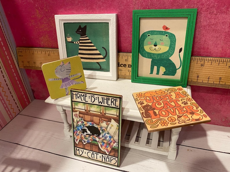 K&ouml;nnte beinhalten: Eine Sammlung von Miniatur-Wandkunst und quadratischen Schildern mit Katzen- und L&ouml;wenillustrationen. Ein Schild tr&auml;gt die Aufschrift "HOME IS WHERE MY CAT NAPS" und ein anderes "JUST A CAT LOVER". Ein Holzlineal dient als Gr&ouml;&szlig;enreferenz.