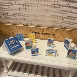 May include: A collection of Hanukkah greeting cards featuring various designs, including a menorah, a cartoon character, and a family. The cards are arranged on a white table with a blue and white gift box that says "Happy Hanukkah".