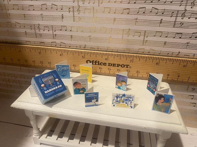 May include: A collection of Hanukkah greeting cards featuring various designs, including a menorah, a cartoon character, and a family. The cards are arranged on a white table with a blue and white gift box that says "Happy Hanukkah".
