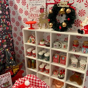 Pode incluir: Uma estante branca com uma variedade de itens tem&aacute;ticos de Natal, incluindo uma coroa, uma casa de gengibre, canecas e jarros. A estante &eacute; decorada com um padr&atilde;o de bengalas de doces vermelhas e brancas.