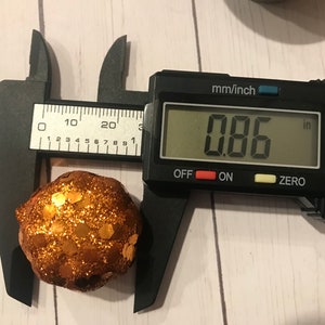 May include: A digital caliper measuring a small, round, orange and gold glitter ball. The caliper reads 0.86 inches.