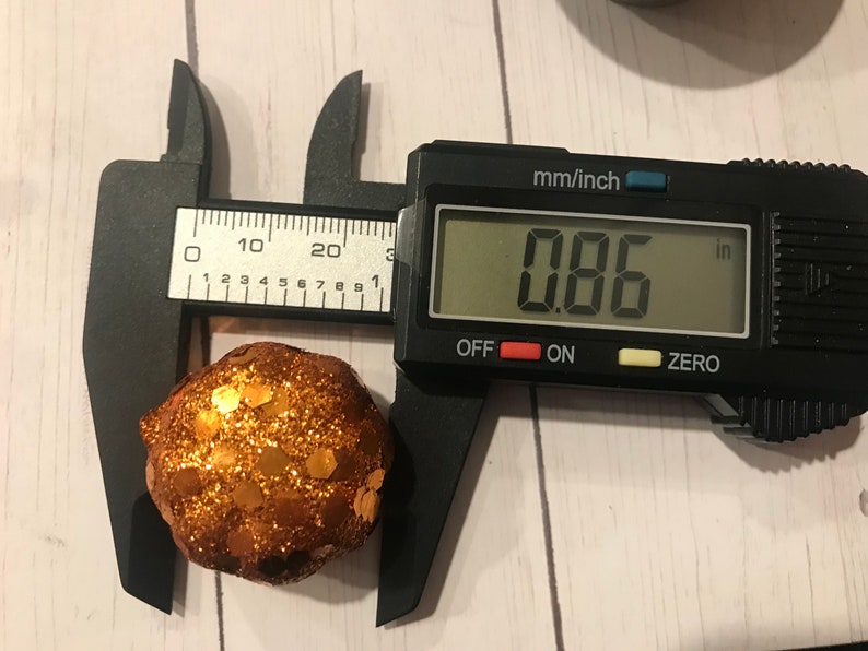 May include: A digital caliper measuring a small, round, orange and gold glitter ball. The caliper reads 0.86 inches.