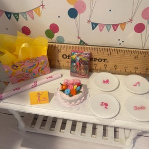 May include: A miniature birthday party scene with a white table. Includes a cake, plates with Barbie logos, a gift bag, a Barbie box, and a rolled-up item with Barbie text.