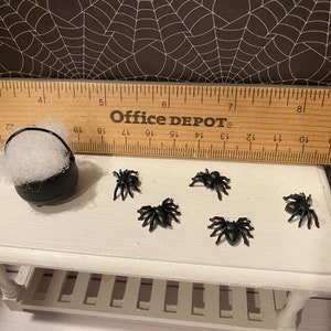 Puede incluir: Un caldero negro lleno de bolas de algod&oacute;n blancas y cinco ara&ntilde;as de pl&aacute;stico negras sobre una mesa blanca con una regla de madera que dice "Office DEPOT".