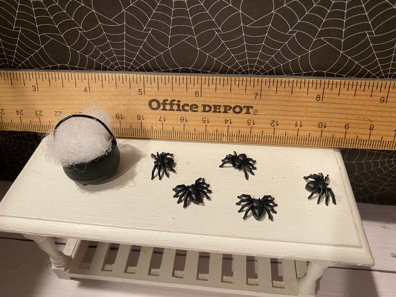 Puede incluir: Un caldero negro lleno de bolas de algod&oacute;n blancas y cinco ara&ntilde;as de pl&aacute;stico negras sobre una mesa blanca con una regla de madera que dice "Office DEPOT".