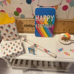 May include: A miniature birthday scene with a blue card reading "HAPPY BIRTHDAY" over a rainbow. Includes a slice of rainbow cake, a box of popcorn, and other decorative items on a white table.