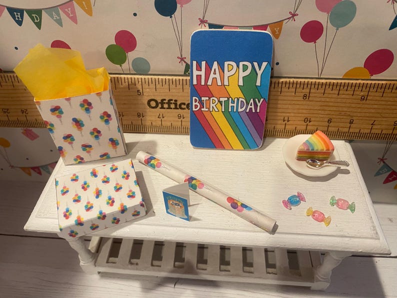 May include: A miniature birthday scene with a blue card reading "HAPPY BIRTHDAY" over a rainbow. Includes a slice of rainbow cake, a box of popcorn, and other decorative items on a white table.