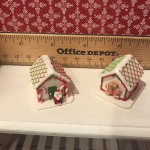 May include: Two miniature gingerbread houses with white icing, red and green accents, and candy cane details. One has a Santa figure, the other a gingerbread man. Festive holiday decor.