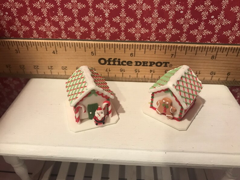 May include: Two miniature gingerbread houses with white icing, red and green accents, and candy cane details. One has a Santa figure, the other a gingerbread man. Festive holiday decor.