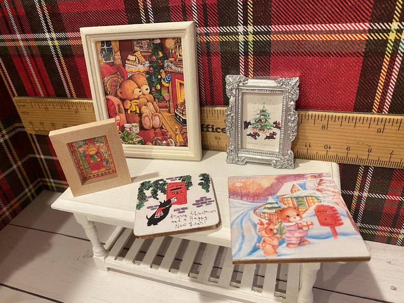 Puede incluir: Una colecci&oacute;n de arte y decoraci&oacute;n navide&ntilde;a en miniatura. Incluye cuadros enmarcados con osos de peluche y un &aacute;rbol de Navidad, y un posavasos con un perro y un buz&oacute;n. Los art&iacute;culos est&aacute;n dispuestos sobre una peque&ntilde;a mesa blanca.