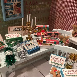 May include: A miniature Christmas scene with a table laden with festive decorations. Boxes of candy canes, Santaland treats, and gingerbread baking mix are arranged alongside a golden candelabra, reindeer figurines, and tinsel. A vintage poster and calendar complete the holiday setting.