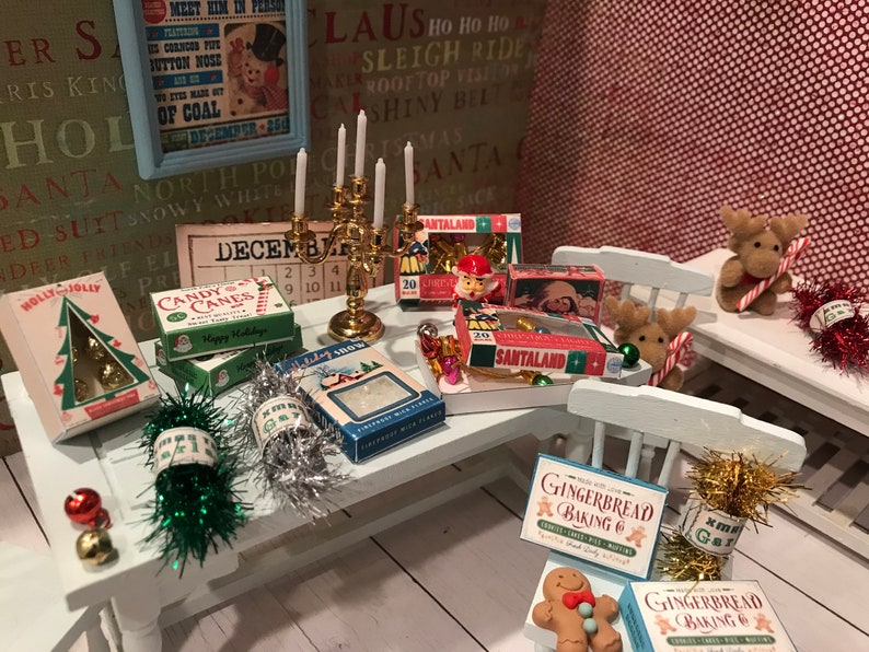May include: A miniature Christmas scene with a table laden with festive decorations. Boxes of candy canes, Santaland treats, and gingerbread baking mix are arranged alongside a golden candelabra, reindeer figurines, and tinsel. A vintage poster and calendar complete the holiday setting.