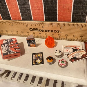 May include: A miniature Halloween scene with a trick or treat box, a Halloween card, a pumpkin, a book, and four Halloween cupcakes. The cupcakes are decorated with a spider, a skull, an eye, and a spider web.