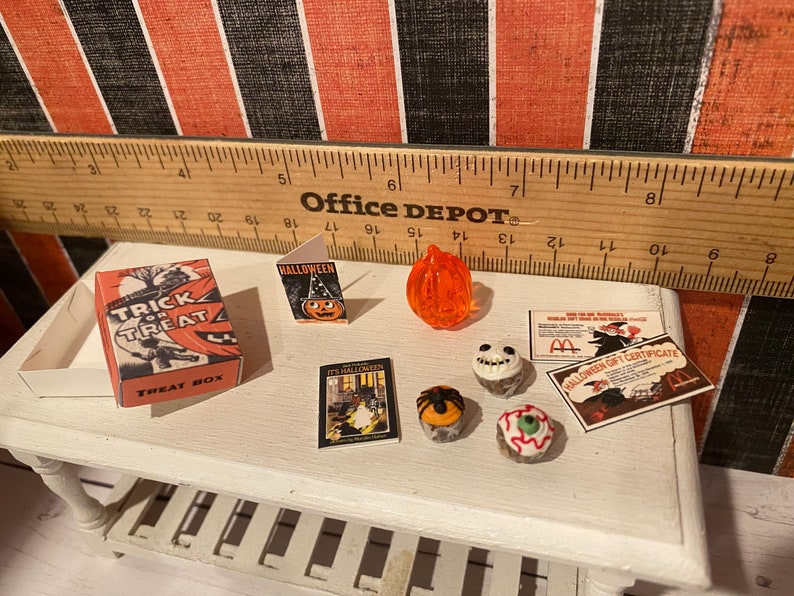 May include: A miniature Halloween scene with a trick or treat box, a Halloween card, a pumpkin, a book, and four Halloween cupcakes. The cupcakes are decorated with a spider, a skull, an eye, and a spider web.