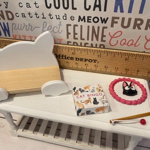 May include: A white miniature cat-shaped bench, a pink and white cat scratching post, a box of cat bingo cards, and a white wooden table with a white wooden toy fishing rod with a red bobber.
