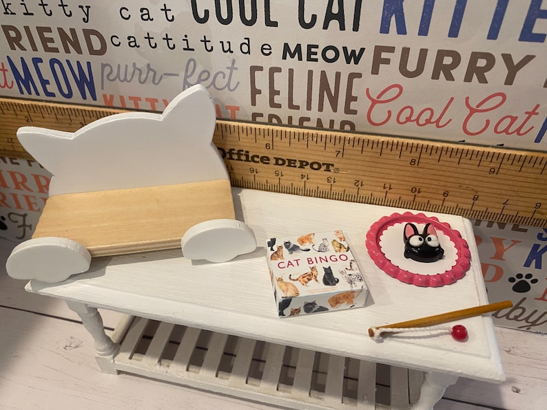 May include: A white miniature cat-shaped bench, a pink and white cat scratching post, a box of cat bingo cards, and a white wooden table with a white wooden toy fishing rod with a red bobber.