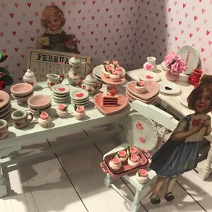 May include: A miniature dollhouse dining set featuring white tables and chairs, decorated with numerous tiny ceramic plates, teacups, and saucers adorned with heart motifs. The display includes miniature cakes, cupcakes with pink roses, and a calendar labeled FEBRUARY.