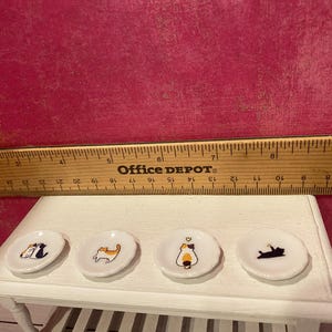 K&ouml;nnte beinhalten: Vier wei&szlig;e Miniatur-Keramikteller mit bunten Katzenillustrationen, die in einer Reihe auf einem wei&szlig;en Tisch angeordnet sind. Ein Holzlineal liegt hinter den Tellern und zeigt einen Durchmesser von etwa 2,5 cm pro Teller.