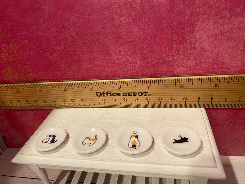 K&ouml;nnte beinhalten: Vier wei&szlig;e Miniatur-Keramikteller mit bunten Katzenillustrationen, die in einer Reihe auf einem wei&szlig;en Tisch angeordnet sind. Ein Holzlineal liegt hinter den Tellern und zeigt einen Durchmesser von etwa 2,5 cm pro Teller.