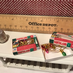 May include: Miniature Christmas lights in boxes labeled "Santaland" with 20 bulbs each. The lights are red, gold, and green. A silver thimble and a wooden ruler are also on the white table. The background is a red and white polka dot pattern.