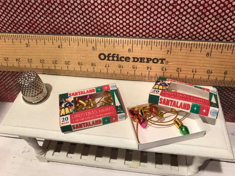 May include: Miniature Christmas lights in boxes labeled "Santaland" with 20 bulbs each. The lights are red, gold, and green. A silver thimble and a wooden ruler are also on the white table. The background is a red and white polka dot pattern.