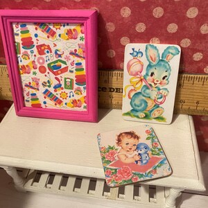 Puede incluir: Una impresi&oacute;n enmarcada en rosa con un colorido patr&oacute;n de juguetes y dos tarjetas de estilo vintage. Una tarjeta presenta un conejito azul con un sonajero, y la otra muestra a un beb&eacute; con un osito de peluche azul. Los art&iacute;culos est&aacute;n en un estante blanco.