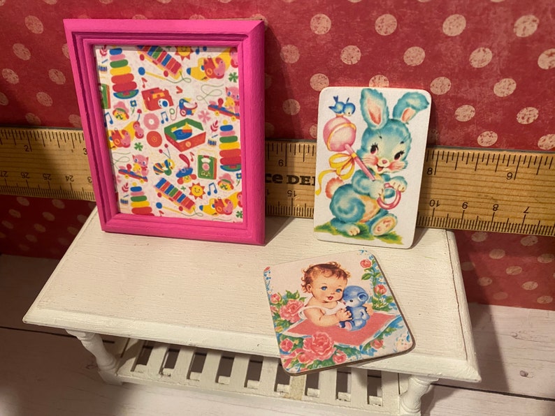 Puede incluir: Una impresi&oacute;n enmarcada en rosa con un colorido patr&oacute;n de juguetes y dos tarjetas de estilo vintage. Una tarjeta presenta un conejito azul con un sonajero, y la otra muestra a un beb&eacute; con un osito de peluche azul. Los art&iacute;culos est&aacute;n en un estante blanco.