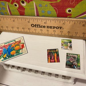 May include: A small white table with a wooden ruler and miniature magazines. The magazines include "Gardening", "Better Homes", and "Living". A "Gardening" sign with colorful flowers is also present.
