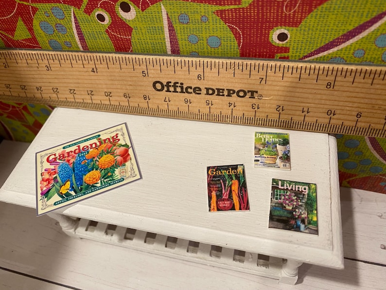 May include: A small white table with a wooden ruler and miniature magazines. The magazines include "Gardening", "Better Homes", and "Living". A "Gardening" sign with colorful flowers is also present.