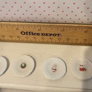 May include: Four small white ceramic plates with different holiday-themed designs. The plates feature a snowman, a wreath, a bunny, and a reindeer.
