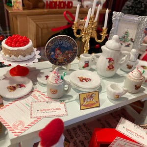 Puede incluir: Una escena navide&ntilde;a en miniatura con una mesa blanca decorada con art&iacute;culos festivos. Incluye un pastel, platos, una tetera, tazas y un marco de fotos. Los art&iacute;culos est&aacute;n decorados con osos de peluche y motivos navide&ntilde;os.