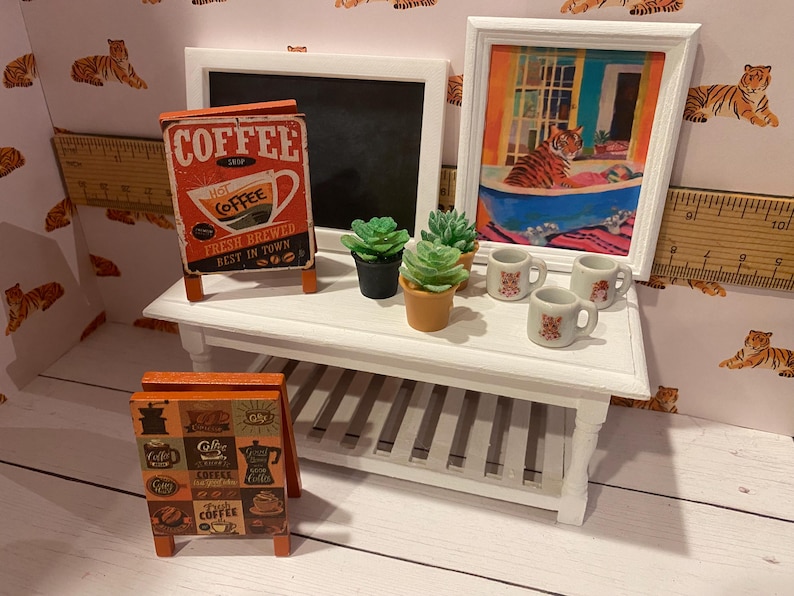 Pode incluir: Uma pequena mesa branca exibe uma imagem emoldurada de um tigre em uma banheira, um quadro-negro e letreiros de caf&eacute;. Pequenas suculentas em vasos e pequenas x&iacute;caras de caf&eacute; tamb&eacute;m est&atilde;o sobre a mesa. O fundo apresenta papel de parede com tema de tigre.