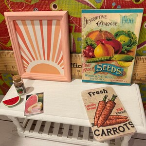 May include: A miniature scene with a framed artwork of a sunburst, a vintage seed catalogue, and a sign for carrots. Also includes a watermelon slice and a small jar. The items are arranged on a white table.
