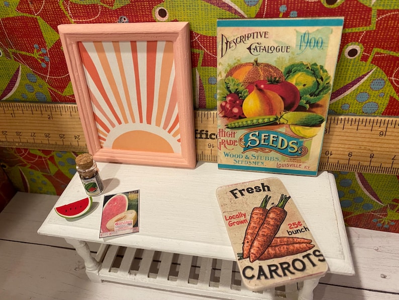 May include: A miniature scene with a framed artwork of a sunburst, a vintage seed catalogue, and a sign for carrots. Also includes a watermelon slice and a small jar. The items are arranged on a white table.