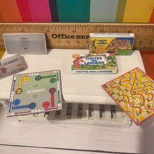 May include: A collection of miniature board games, including "Sorry!" and "Chutes and Ladders", are arranged on a white surface. The games feature colourful boards and boxes. The "Sorry!" board game is approximately 10 cm square.
