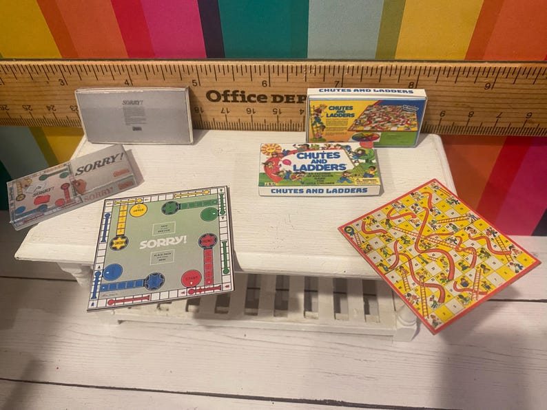 May include: A collection of miniature board games, including "Sorry!" and "Chutes and Ladders", are arranged on a white surface. The games feature colourful boards and boxes. The "Sorry!" board game is approximately 10 cm square.