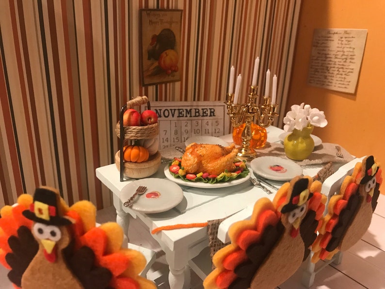 May include: A miniature Thanksgiving dinner scene with a roasted turkey, plates, and a candle holder. The table is set with felt turkey decorations, a calendar showing November, and a tiered stand with apples and a pumpkin. The background has striped wallpaper.