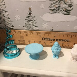 May include: A set of four miniature Christmas decorations, including a blue spiral Christmas tree, a blue table, a blue Santa Claus figurine, and a white Santa Claus figurine. The decorations are arranged on a white table with a wooden ruler behind them. The ruler reads "Office DEPOT&reg;".