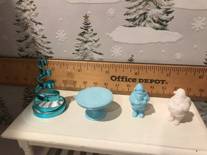 May include: A set of four miniature Christmas decorations, including a blue spiral Christmas tree, a blue table, a blue Santa Claus figurine, and a white Santa Claus figurine. The decorations are arranged on a white table with a wooden ruler behind them. The ruler reads "Office DEPOT&reg;".