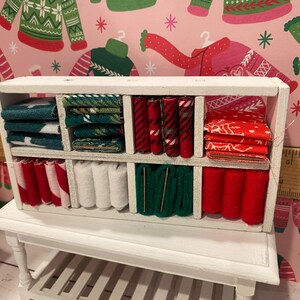 May include: A miniature white wooden shelf unit filled with various fabrics. The fabrics are in shades of red, green, and white, with patterns including stripes and holiday designs. The shelf sits on a small white table.
