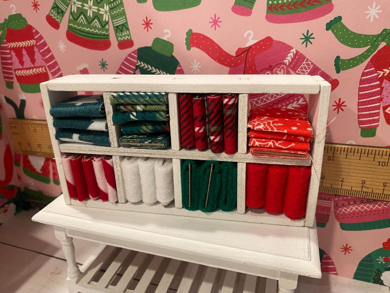 May include: A miniature white wooden shelf unit filled with various fabrics. The fabrics are in shades of red, green, and white, with patterns including stripes and holiday designs. The shelf sits on a small white table.
