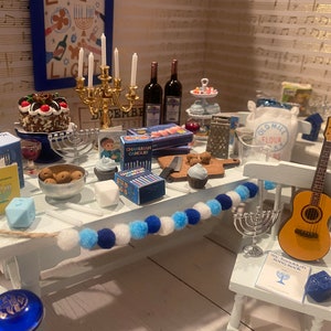 May include: A miniature Hanukkah scene with a white table set for a festive celebration. The table is decorated with a blue and white pom-pom garland, a silver menorah, and a variety of blue and white decorations. There is a chocolate cake with a blue and white star on top, a small guitar, and a bag of flour labeled "Old Mill Flour".