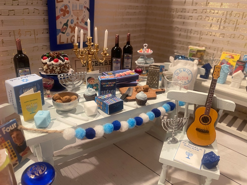 May include: A miniature Hanukkah scene with a white table set for a festive celebration. The table is decorated with a blue and white pom-pom garland, a silver menorah, and a variety of blue and white decorations. There is a chocolate cake with a blue and white star on top, a small guitar, and a bag of flour labeled "Old Mill Flour".