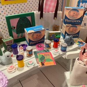 May include: Miniature baby items including Pampers boxes, Gerber Rice boxes, a duck, bottles, and a book titled "Chicka Chicka Boom Boom" are arranged on a white surface. A framed turtle picture and a blue elephant figurine are also present.