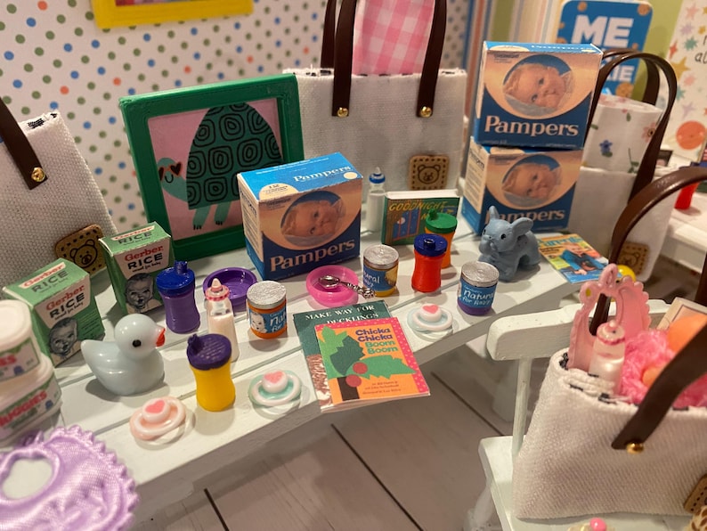 May include: Miniature baby items including Pampers boxes, Gerber Rice boxes, a duck, bottles, and a book titled "Chicka Chicka Boom Boom" are arranged on a white surface. A framed turtle picture and a blue elephant figurine are also present.