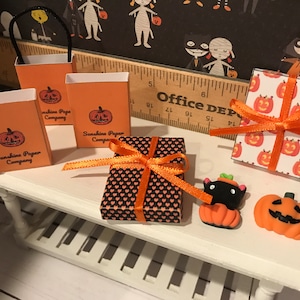 Puede incluir: Un expositor de mesa con bolsas de regalo y cajas con tem&aacute;tica de Halloween. Tres bolsas de regalo naranjas con dise&ntilde;os de calabazas negras y el texto "Sunshine Paper Company" est&aacute;n sobre la mesa. Una caja de regalo negra y naranja con un patr&oacute;n de coraz&oacute;n est&aacute; atada con una cinta naranja. Una caja de regalo blanca con calabazas naranjas est&aacute; atada con una cinta naranja. Una figurita de gato negro con orejas de calabaza naranja est&aacute; sentada sobre una peque&ntilde;a calabaza naranja. Una calabaza naranja m&aacute;s grande con una cara negra est&aacute; sobre la mesa.
