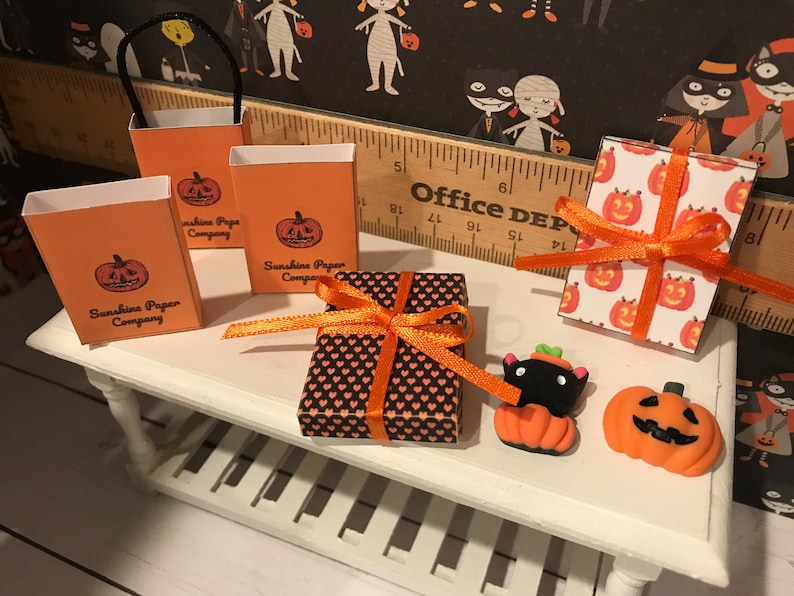 Puede incluir: Un expositor de mesa con bolsas de regalo y cajas con tem&aacute;tica de Halloween. Tres bolsas de regalo naranjas con dise&ntilde;os de calabazas negras y el texto "Sunshine Paper Company" est&aacute;n sobre la mesa. Una caja de regalo negra y naranja con un patr&oacute;n de coraz&oacute;n est&aacute; atada con una cinta naranja. Una caja de regalo blanca con calabazas naranjas est&aacute; atada con una cinta naranja. Una figurita de gato negro con orejas de calabaza naranja est&aacute; sentada sobre una peque&ntilde;a calabaza naranja. Una calabaza naranja m&aacute;s grande con una cara negra est&aacute; sobre la mesa.